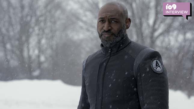 Adrian Holmes as Admiral Robert April in Star Trek: Strange New Worlds. He's wearing a black overcoat with the Starfleet Delta insignia worn as a patch on its left arm, as he stands in snowfall against the backdrop of a snow-covered forest.