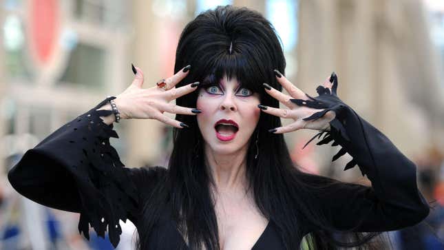 Elvira (horror-host alter ego of Cassandra Peterson) poses with her hands on either side of her shocked face while wearing a black gown and wig.
