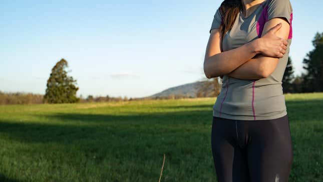 A woman in workout gear hugs herself as if for warmth while outside in nature on a sunny day