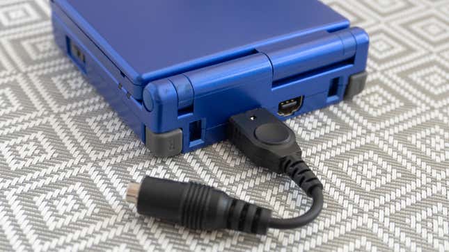 A close-up of a headphone adapter connected to a blue GBA SP against a patterned background.