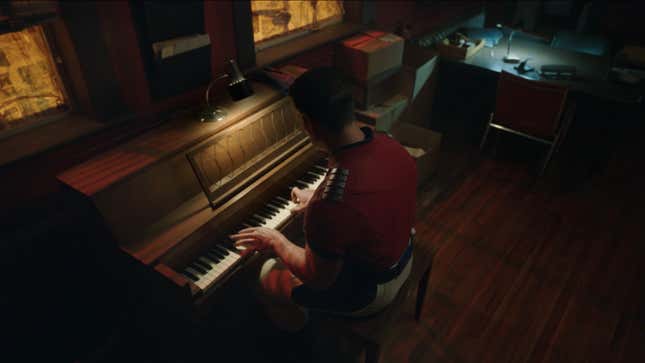 Peacemaker somberly plays an upright piano in a darkened room.