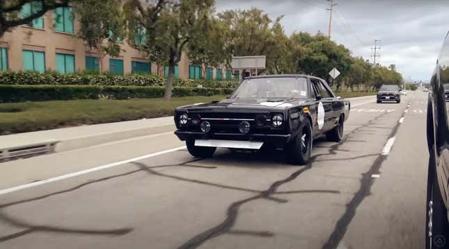 A black 1967 Plymouth GTX drives on a road with tar snakes.