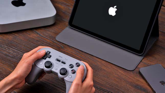 A brace  of hands holding the 8BitDo Pro 2 controller implicit    a woody  table  with an iPad, iPhone, and Mac sitting nearby.