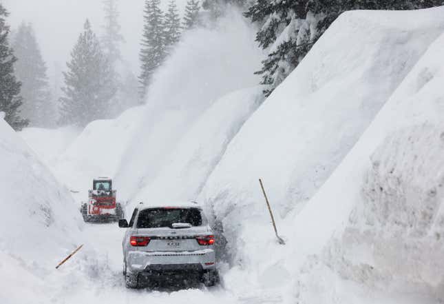 A car navigates behind a snowblower through piles of drifts from a season of intense snowfall in Mammoth Lakes, CA. 
