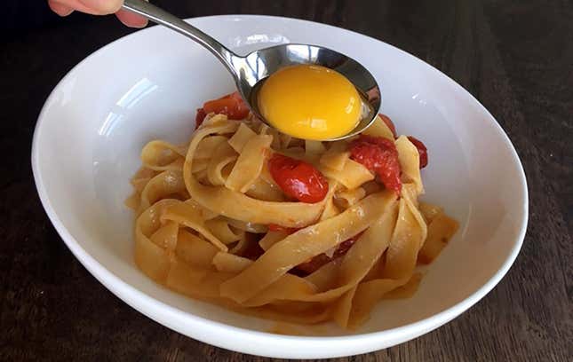 Egg yolk being spooned onto a bowl of pappardelle and tomatoes