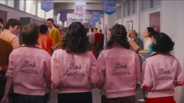 Four teenage girls wearing pink jackets with the words "Pink Ladies" stitched on the back walk away from the camera down a crowded high school hallway