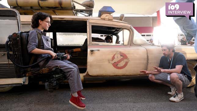 Mckenna Grace in costume as Phoebe in Ghostbusters: Afterlife sits in the ecto 1 gunner seat as director Jason Reitman squats in front of her.