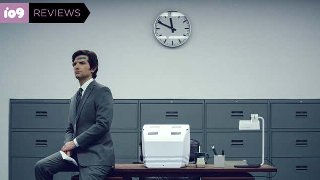 Adam Scott sits stiffly on a desk, wearing a grey suit and a blue bandage on his forehead, in a scene from Severance.