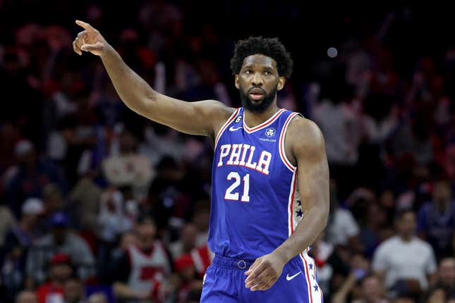 A Black man with a beard, wearing a blue 76ers uniform with white and red trim, points to something on the floor of an NBA game. The crowd is seen in the background.