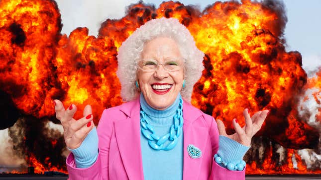 A smiling old woman stands in front of plumes of flames.