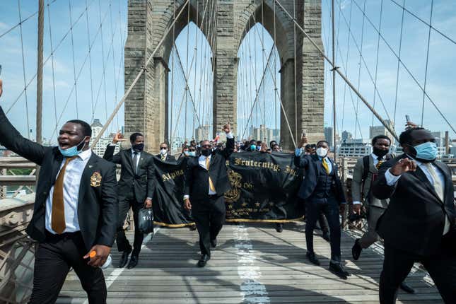 A group of protesters from the Alpha Phi Alpha fraternity holding up fists in the air and wearing suits walk with the arches of the Brooklyn Bridge behind them as they walk across the Brooklyn Bridge in protest of police racism. 