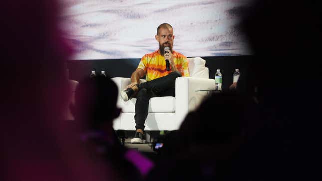 Jack Dorsey sitting in a white armchair holding a microphone bordered on both sides by black silhouettes.