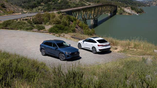 A blue and a white 2024 Audi Q8 E-Tron SUVs, one a Sportback, are parked in gravel near a bridge.