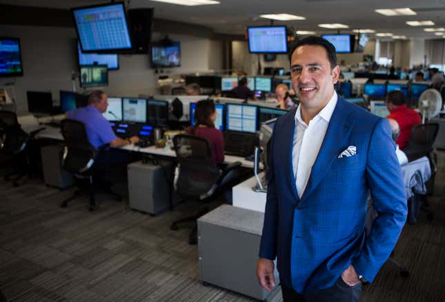 A Latino man with dark hair and a bright blue jacket with a pocket square and no tie stands in a room full of men and women working at computers.
