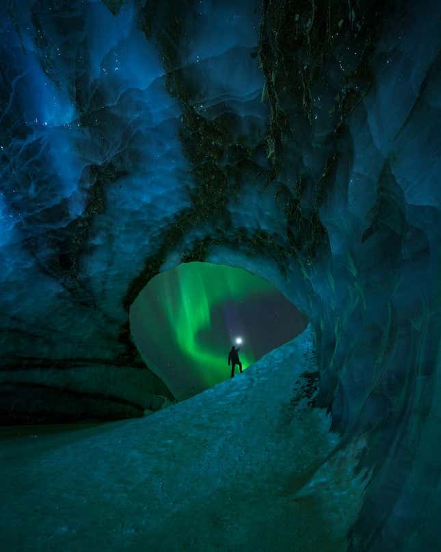 A person silhouetted under the Northern Lights, looking out from an ice cave.
