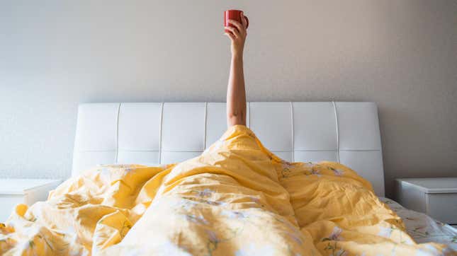 Person covered in blanket waking up, holding mug of coffee