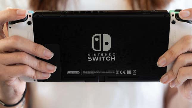 A woman's hands are seen holding a black and white Nintendo Switch OLED model, viewed from the back with the logo visible