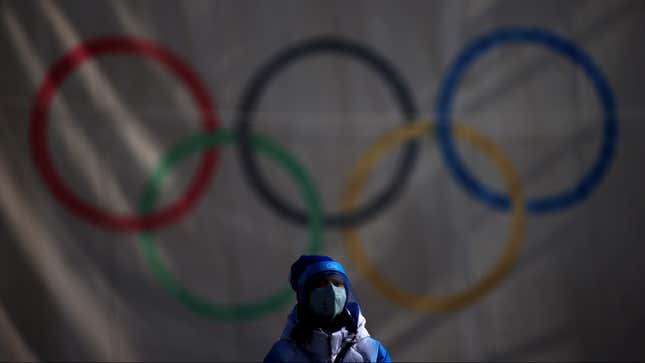 A person in winter clothes stands in front of a flag of the Olympic rings wearing a mask and face shield.
