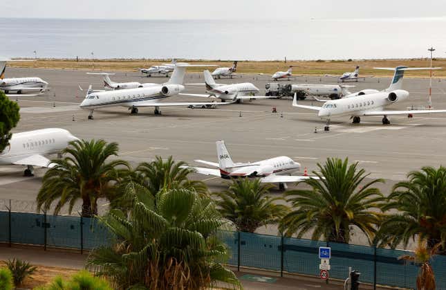 Private jets are parked on the tarmac. Behind them is the ocean, in the foreground are trees with palm fronds.