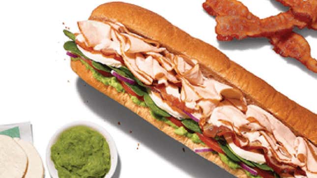 Subway's turkey cali sub with a side of green sauce and three slices of bacon on white background