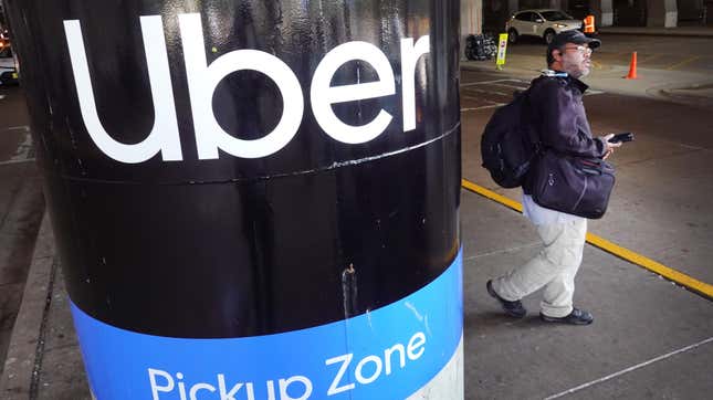 A Pole with the Uber logo and text under it saying "pickup zone." A man with a bag walks by holding his phone, waiting for his Uber pickup.