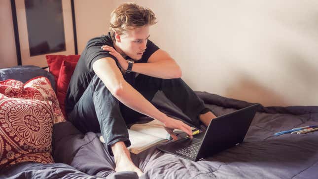 Young man sitting on bed, working on laptop with textbook open