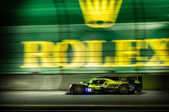 A neon yellow and black prototype racecar drives on a racetrack in front of a large Rolex banner at night.
