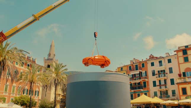 A completely orange Fiat -- tires and all -- is held aloft by a crane above what appears to be a large gray vat of paint. It's surrounded by palm trees and colorful buildings denoting a tropical local.