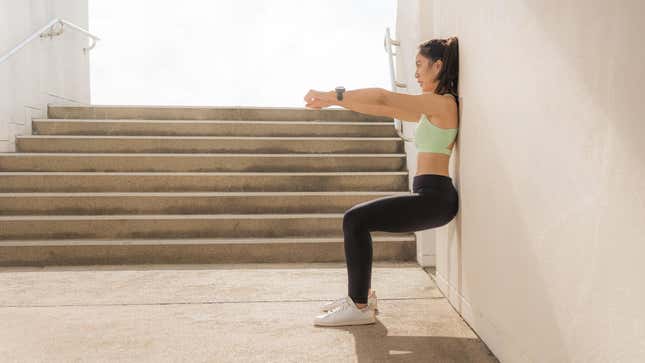 A woman in workout clothes does a wall sit exercise against a stone wall