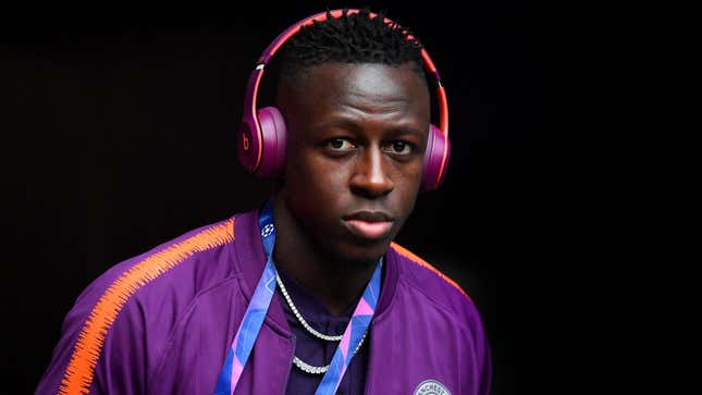Benjamin Mendy stands in front of a black background wearing orange and purple Beats headphones that match his team jacket.