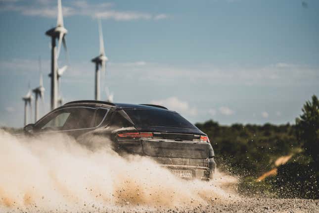 an action photo of a 2024 porsche macan electric prototype. we see the rear of the vehicle as it power-slides across a dirt road, kicking up dust. the vehicle is painted black and wearing vinyl wrapping to disguise its design.