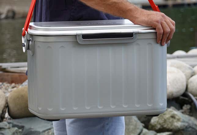 A person carrying the Oyster Tempo cooler using its red carrying strap near a small river.