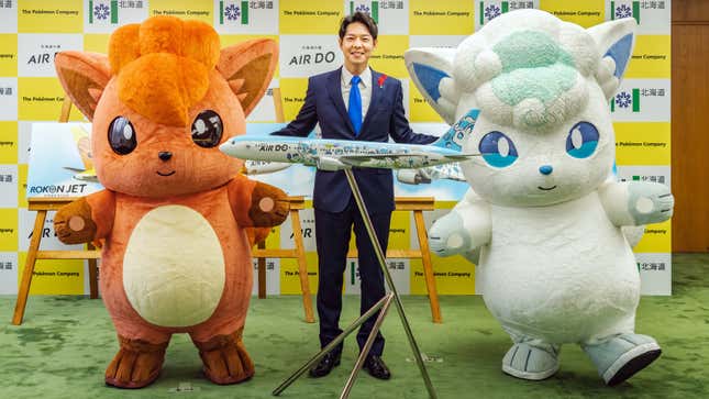 A man in a suit shows off a model plane version of the Vulpix plane and is flanked by two people dressed as  Vulpix. 