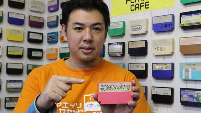 Junji Seki holds and points to a pink Famicom cartridge with someone's name on it. A wall of signed cartridges is behind him.