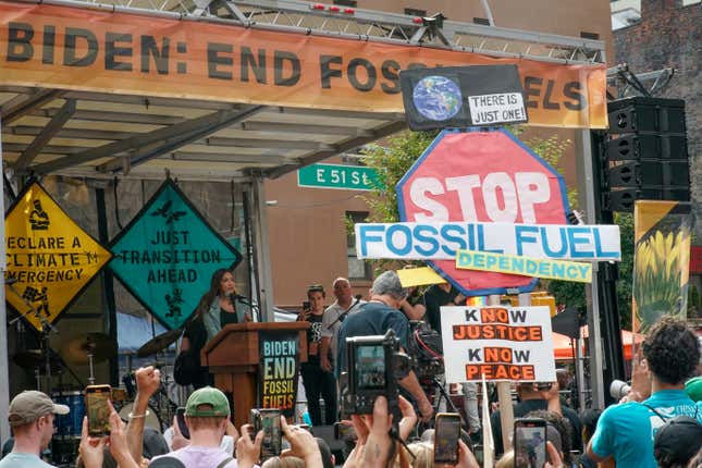 Rep. Alexandria Ocasio-Cortez speaks on a stage with a banner reading "Biden: End fossil fuels now" in front of a crowd holding signs in New York City.