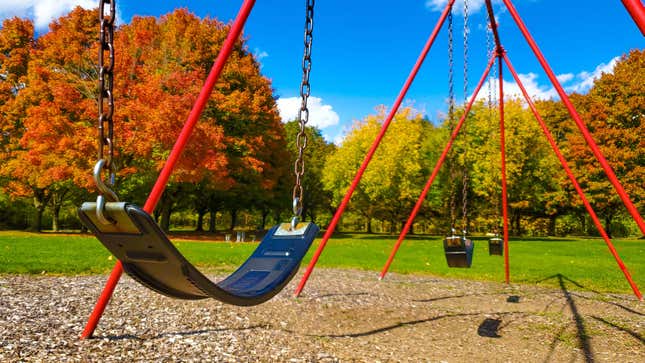An empty playground swing set, pictured on a beautiful blue sky day in a large tree-dotted park in the fall