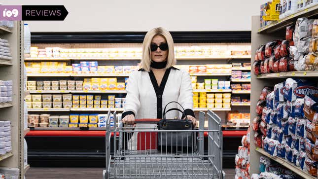 A blonde woman wearing dramatic sunglasses and a black and white blouse pushes a supermarket shopping cart in Jakob's Wife.