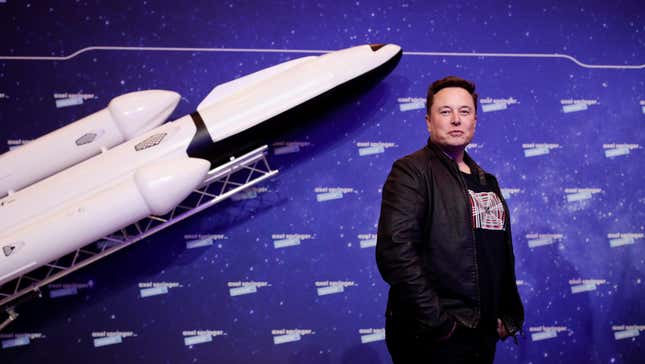 Elon Musk is photographed with a white miniature rocket in the background.