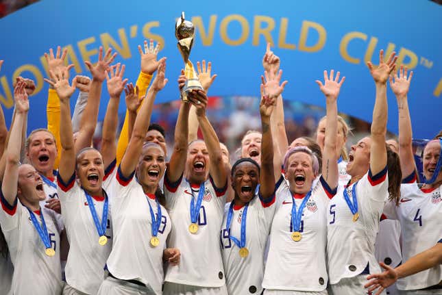 The US women's national soccer team raise their hands and shout in victory, Carli Lloyd in the center lifting the trophy.