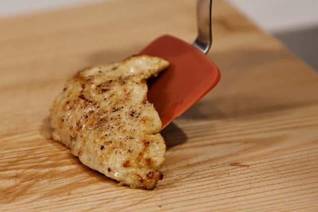 A cooked piece of cultivated chicken breast being laid on a wooden cutting board with a reddish-orange spatula.