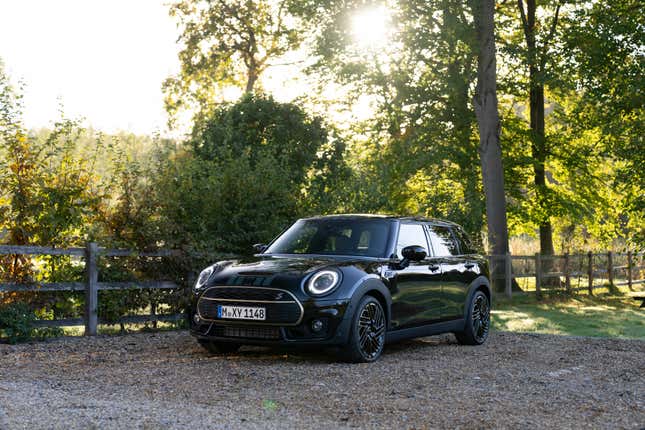 A black and gold 2023 Mini Cooper Clubman is parked on gravel in front of some trees.