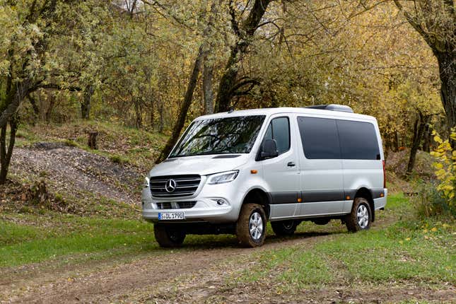 a silver 2023 mercedes benz sprinter awd van drives down a dirt path on an overcast day. there are trees in the background with yellow leaves, indicating late autumn in germany.