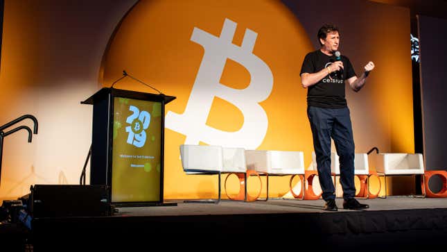 Alex Mashinsky stands on a stage with a microphone in his hand wearing a shirt reading "Celsius." Behind him are several empty chairs and a giant Bitcoin symbol.