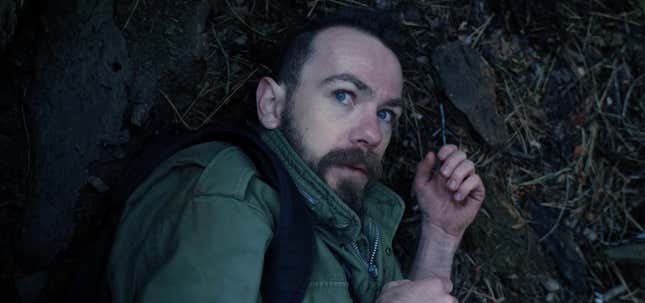 A man in a green Army jacket cowers on the forest floor.