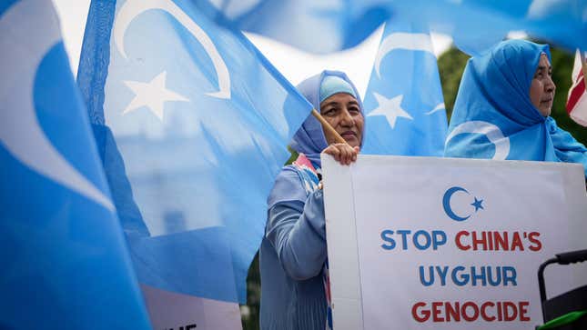 A woman in a blue hijab holds a sign reading Stop China's Uyghur Genocide behind blue flags representing East Turkestan.
