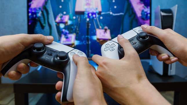 Two sets of hands hold PS5 controllers with a TV visible in the background displaying a competitive game