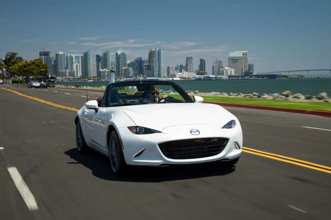 A white 2023 Mazda Miata drives along a waterfront with its top down.