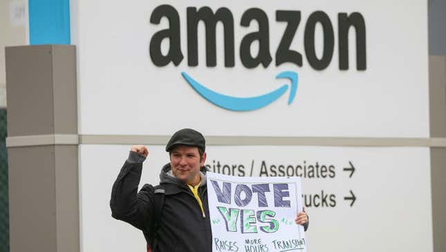 an amazon labour union organizer holding a sign that says "vote yes" stands in front of an amazon warehouse sign, with his fist raised