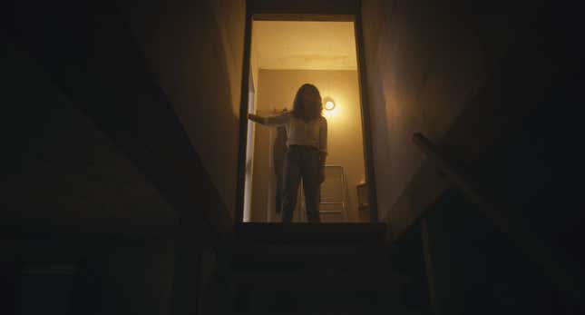 A woman stands at the top of a dark staircase with a light behind her.