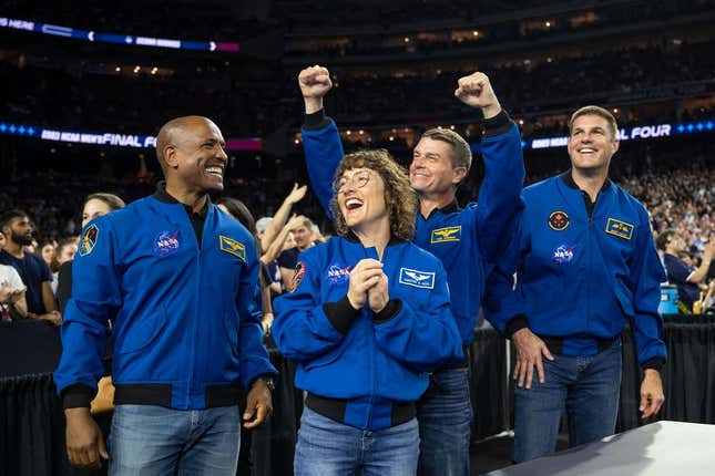 Four astronauts in jeans and blue jackets, looking delighted at a basketball game.